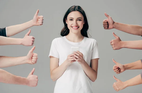 Vista parcial de hombres y mujeres mostrando los pulgares hacia arriba cerca de chica alegre con teléfono inteligente aislado en gris - foto de stock