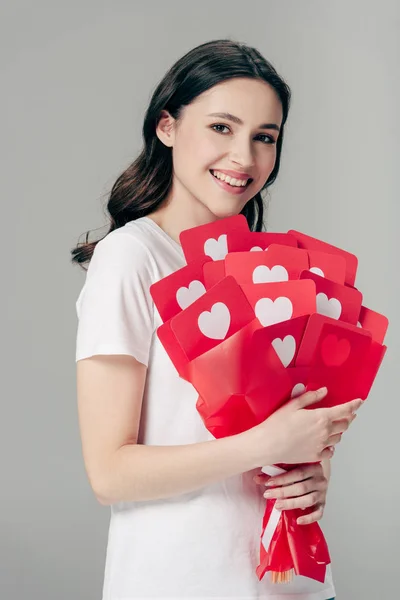 Feliz chica bonita sosteniendo ramo de tarjetas de corte de papel rojo con símbolos de corazones y mirando a la cámara aislada en gris - foto de stock