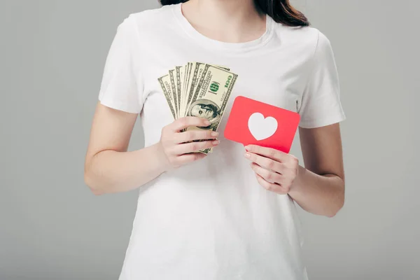 Частичный вид молодой девушки с банкнотами в долларах и красной карточкой с символом сердца, изолированным на сером — стоковое фото