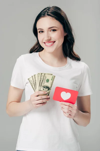 Hermosa chica sonriente sosteniendo billetes de dólar y tarjeta de corte de papel rojo con símbolo del corazón aislado en gris - foto de stock