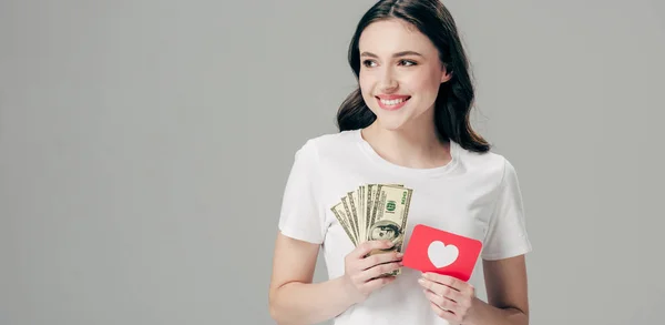 Panoramic shot of happy girl holding dollar banknotes and red paper cut card with heart symbol isolated on grey — Stock Photo