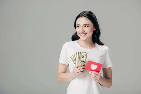 Hermosa chica sonriente sosteniendo billetes de dólar y tarjeta de corte de papel rojo con símbolo del corazón aislado en gris - foto de stock