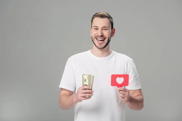 Joyeux homme en t-shirt blanc tenant billets en dollars et carte de papier rouge avec symbole de coeur isolé sur gris — Photo de stock