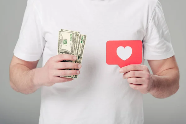 Частичный вид человека, держащего банкноты по доллару и красную карточку с символом сердца, изолированным на сером — стоковое фото