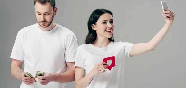 Panoramaaufnahme eines lächelnden Mädchens, das eine rote Karte mit Herzsymbol in der Hand hält und ein Selfie mit dem Smartphone macht, in der Nähe eines gutaussehenden Mannes, der Dollarnoten isoliert auf grau zählt — Stockfoto