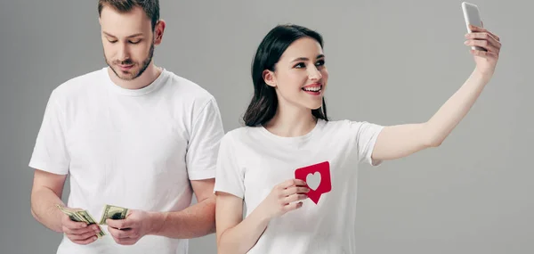 Plano panorámico de niña sonriente sosteniendo tarjeta de corte de papel rojo con símbolo del corazón y haciendo selfie con teléfono inteligente cerca de hombre guapo contando billetes de dólar aislados en gris - foto de stock