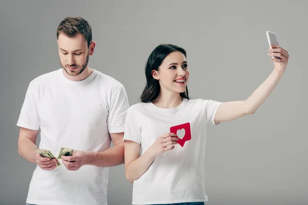 Alegre chica sosteniendo papel rojo tarjeta de corte con símbolo del corazón y haciendo selfie con teléfono inteligente cerca de hombre guapo contando billetes de dólar aislados en gris - foto de stock