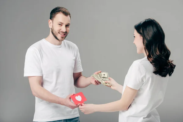 Homem bonito sorrindo dando notas de dólar para a jovem com cartão de corte de papel vermelho com símbolo de coração no fundo cinza — Fotografia de Stock