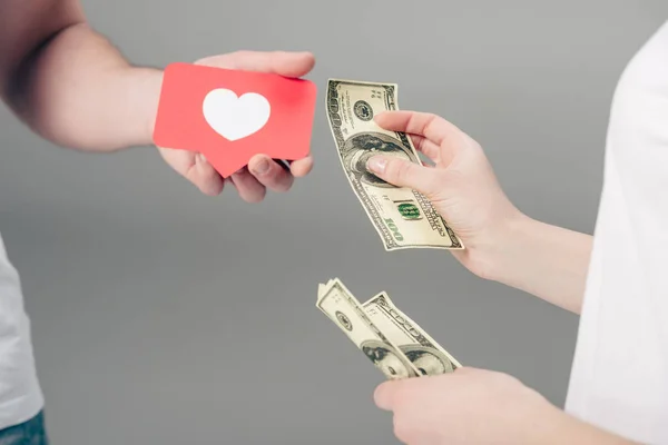 Vista recortada de la mujer dando billetes de dólar al hombre con tarjeta de corte de papel rojo con símbolo del corazón sobre fondo gris - foto de stock