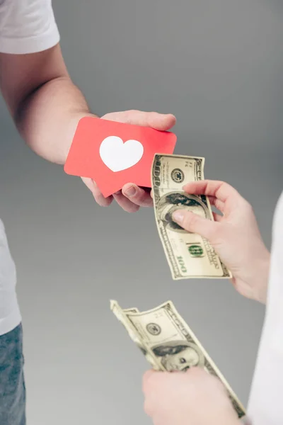 Partial view of woman giving dollar banknotes to man with red paper cut card with heart symbol on grey background — Stock Photo