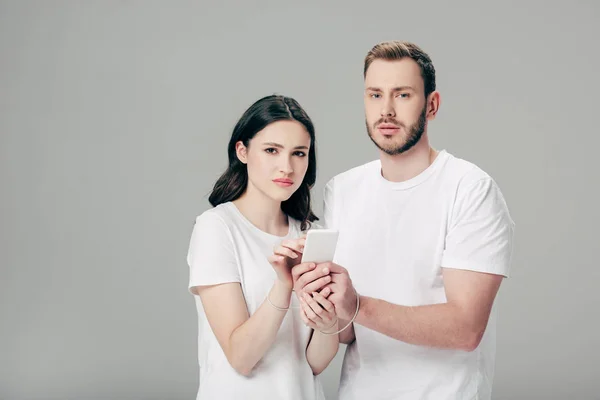 Giovane uomo e donna in t-shirt bianche con cavo usb intorno alle mani utilizzando smartphone isolato su grigio — Foto stock