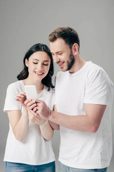 Веселий чоловік і жінка в білих футболках з USB-кабелем навколо рук, використовуючи смартфон ізольовано на сірому — стокове фото