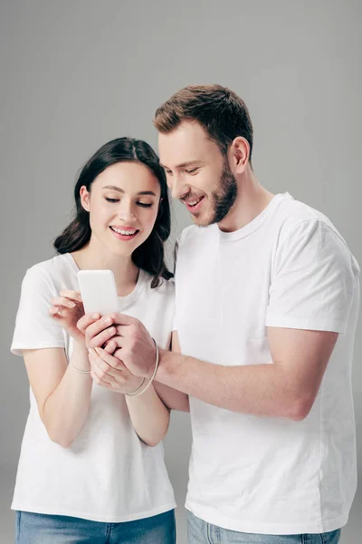 Fröhliche Mann und Frau in weißen T-Shirts mit USB-Kabel um die Hände mit Smartphone isoliert auf grau — Stockfoto