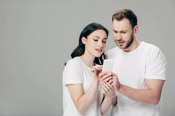 Focalisé homme et femme en t-shirts blancs avec câble USB autour des mains en utilisant smartphone isolé sur gris — Photo de stock