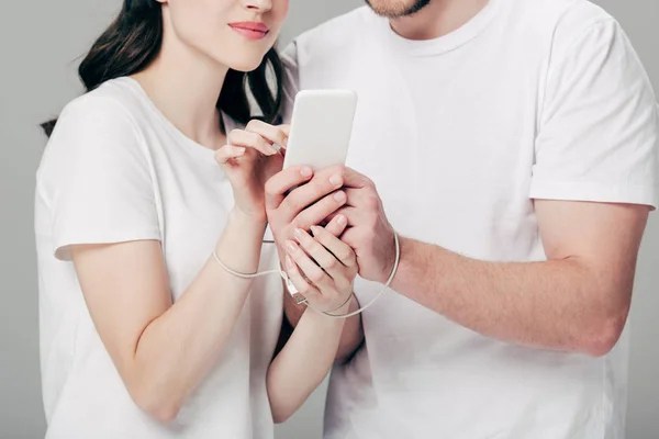 Vista parcial del hombre y la mujer con cable USB alrededor de las manos utilizando el teléfono inteligente aislado en gris - foto de stock