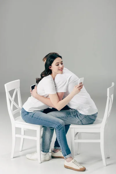 Joven pareja abrazando mientras se sienta en sillas blancas y el uso de teléfonos inteligentes sobre fondo gris - foto de stock