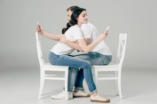 Vue latérale du jeune couple embrassant assis sur des chaises blanches et utilisant des smartphones sur fond gris — Photo de stock