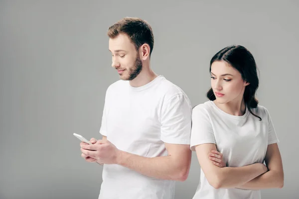 Ofendido chica bonita de pie con las manos cruzadas cerca novio guapo usando teléfono inteligente aislado en gris - foto de stock