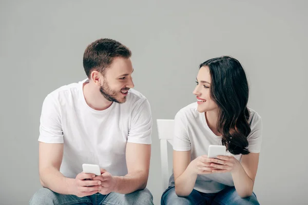 Coppia sorridente guardarsi mentre si siede sulle sedie e utilizzando smartphone isolato su grigio — Foto stock