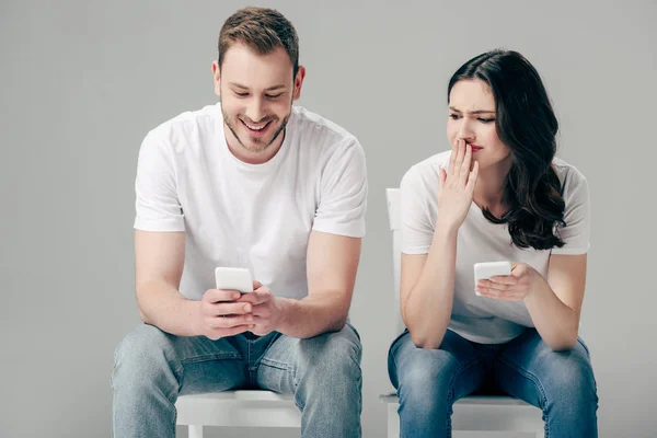 Shocked girl looking at boyfriend using smartphone isolated on grey — Stock Photo