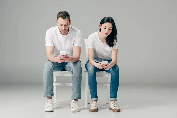 Концентрированные мужчина и женщина в белых футболках и синих джинсах, сидящих на стульях и использующих смартфоны на сером фоне — стоковое фото