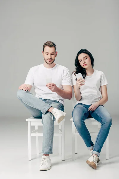 Hombre y mujer enfocados en camisetas blancas y vaqueros azules sentados en sillas y usando teléfonos inteligentes sobre fondo gris - foto de stock