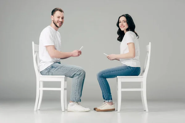 Усміхнений чоловік і жінка в білих футболках і синіх джинсах, сидячи на стільцях і використовуючи смартфони на сірому фоні — стокове фото