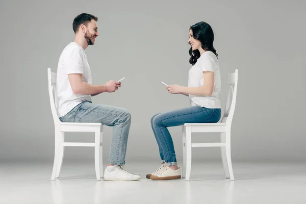 Vista lateral de homem e mulher sorridente em camisetas brancas e jeans azuis sentados em cadeiras com smartphones e olhando um para o outro em fundo cinza — Fotografia de Stock