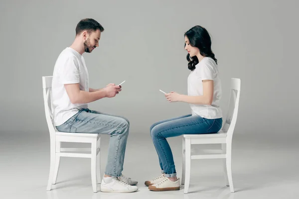 Side view of concentrated man and woman in white t-shirts and blue jeans using smartphones while sitting on chairs on grey background — Stock Photo