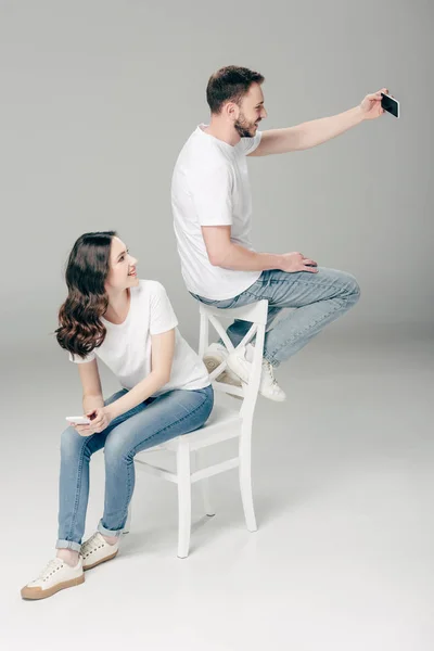 Chica sonriente mirando sentado en la silla y mirando al hombre alegre tomando selfie con teléfono inteligente mientras está sentado en el respaldo de la silla sobre fondo gris - foto de stock