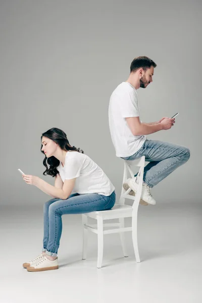 Bonita mujer joven en silla y hombre guapo en el respaldo de la silla usando teléfonos inteligentes sobre fondo gris - foto de stock