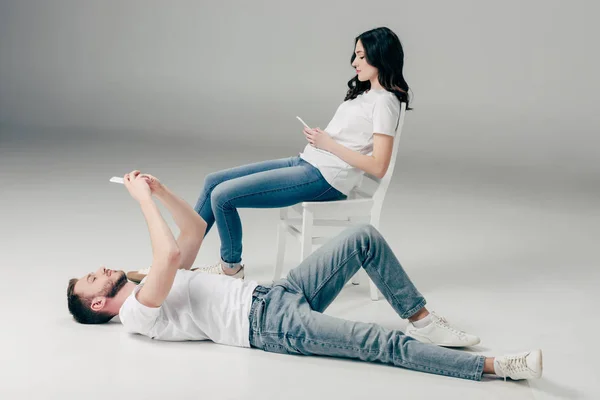 Konzentrierte junge Frau sitzt auf Stuhl und benutzt Smartphone neben gutaussehendem Mann, der auf dem Boden liegt und Selfie auf grauem Hintergrund macht — Stockfoto