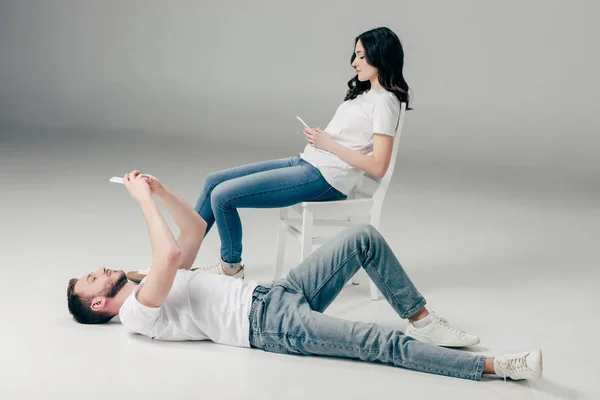 Concentrated young woman sitting on chair and using smartphone near handsome man lying on floor and taking selfie on grey background — Stock Photo