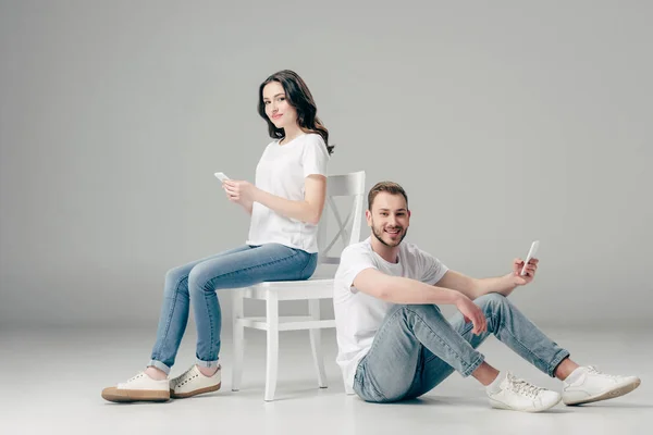 Усміхнена жінка використовує смартфон біля веселого чоловіка, що сидить на підлозі з смартфоном на сірому фоні — стокове фото
