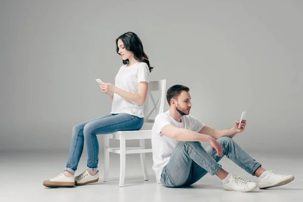 Mujer concentrada usando teléfono inteligente cerca de hombre enfocado sentado en el suelo con teléfono inteligente sobre fondo gris - foto de stock