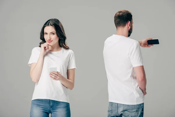 Sorrindo menina atraente em t-shirt branca de pé perto jovem tomando selfie com smartphone isolado em cinza — Fotografia de Stock