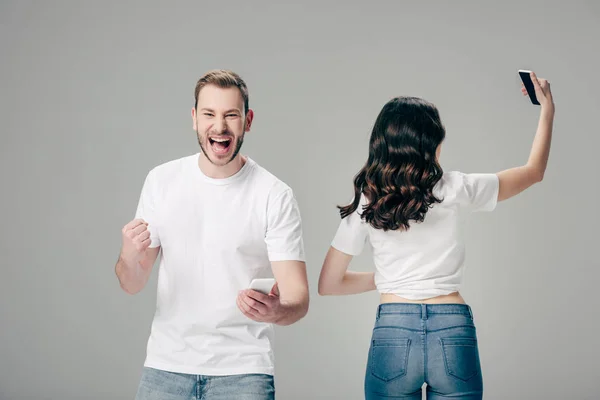 Excited young man with smartphone showing winner gesture while standing near girl taking selfie isolated on grey — Stock Photo