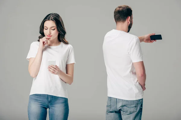 Muchacha joven pensativa usando el teléfono inteligente, mientras que de pie cerca de hombre tomando selfie aislado en gris - foto de stock