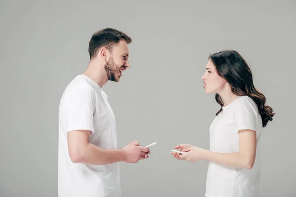 Vista lateral de pareja joven en camisetas blancas peleando mientras sostiene teléfonos inteligentes aislados en gris - foto de stock
