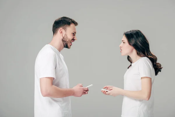 Vista lateral do jovem casal em camisetas brancas brigando enquanto segurando smartphones isolados em cinza — Fotografia de Stock