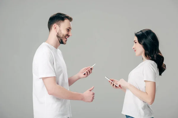 Hombre guapo mostrando el pulgar hacia arriba mientras está de pie cerca de chica bonita con teléfono inteligente aislado en gris - foto de stock