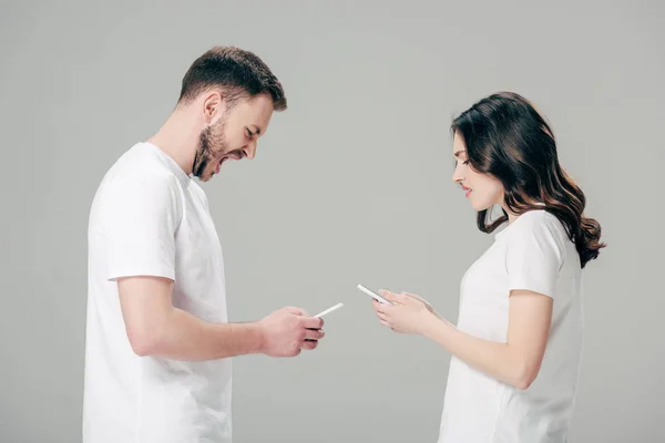 Hombre guapo usando teléfono inteligente y gritando de pie cerca de chica disgustado con teléfono inteligente aislado en gris - foto de stock