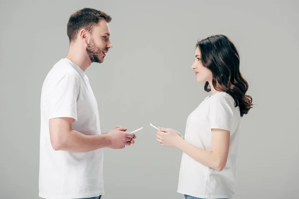 Young man and woman in white t-shirts looking at each other while holding smartphones isolated on grey — Stock Photo