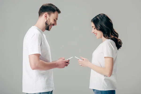 Vista lateral de sonriente joven hombre y mujer en camisetas blancas usando teléfonos inteligentes aislados en gris - foto de stock