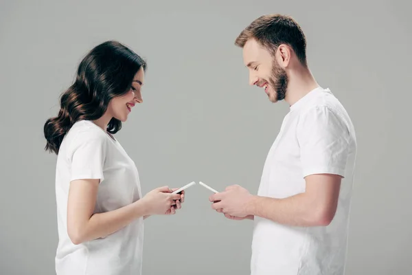 Вид сбоку веселый мужчина и женщина в белых футболках с использованием смартфонов, изолированных на сером — стоковое фото