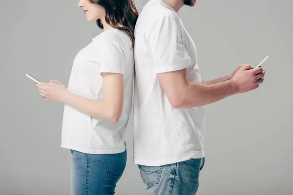 Cropped view of cheerful man and woman in white t-shirts standing back to back and using smartphones isolated on grey — Stock Photo