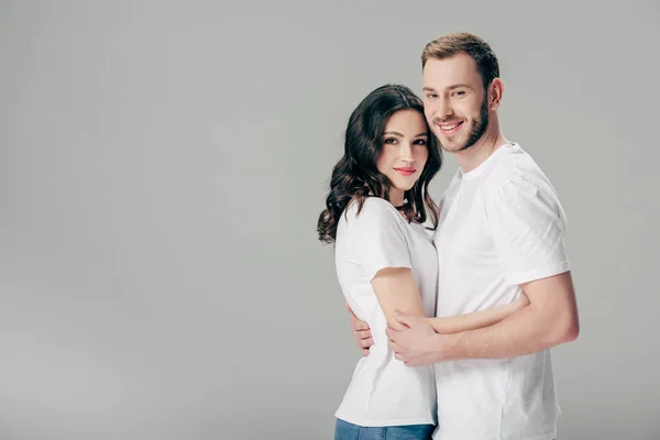 Jeune couple souriant en t-shirts blancs embrassant et regardant la caméra isolée sur gris — Photo de stock