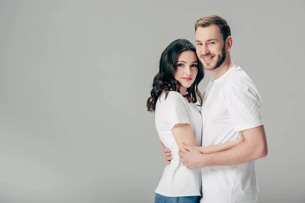 Smiling young couple in white t-shirts embracing and looking at camera isolated on grey — Stock Photo