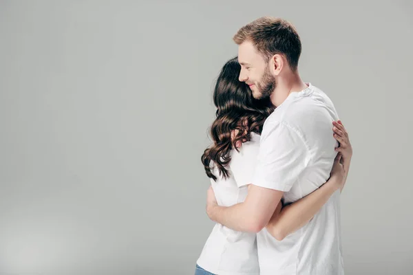 Glückliches junges Paar in weißen T-Shirts, die sich auf grauem Hintergrund umarmen — Stockfoto