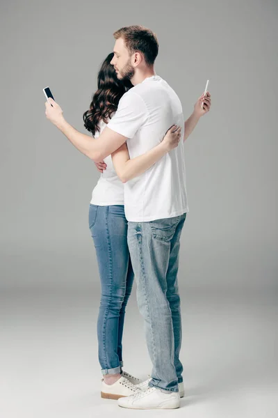 Молода пара в білих футболках і синіх джинсах, що обіймаються при використанні смартфонів на сірому фоні — стокове фото